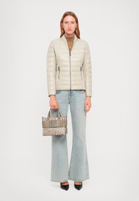 Woman wearing a light beige quilted jacket, brown turtleneck, light blue flared jeans, black pointed shoes, holding a beige patterned handbag.