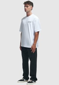 Hvit bomull t-skjorte med korte ermer og en liten svart logo på brystet, sammen med svarte denimbukser og svarte sneakers.