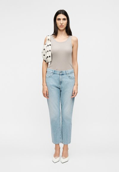 Γυναίκα που στέκεται, φοράει γκρι τοπ χωρίς μανίκια, ανοιχτόχρωμο μπλε τζιν, λευκά μυτερά τακούνια και μια λευκή τσάντα με μαύρα κυκλικά σχέδια στον ώμο της.