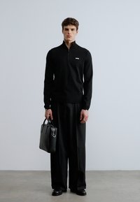 Svart genser med glidelås ved halsen og teksturert stoff, kombinert med vide svarte bukser. Modellen holder en svart skinnveske med sølvdetalj.