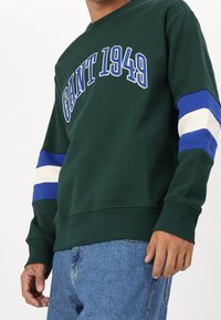 Sweatshirt vert avec le texte "GANT 1949" en bleu, présentant des rayures contrastantes bleu et blanc sur les manches. Poignets et ourlet côtelés.