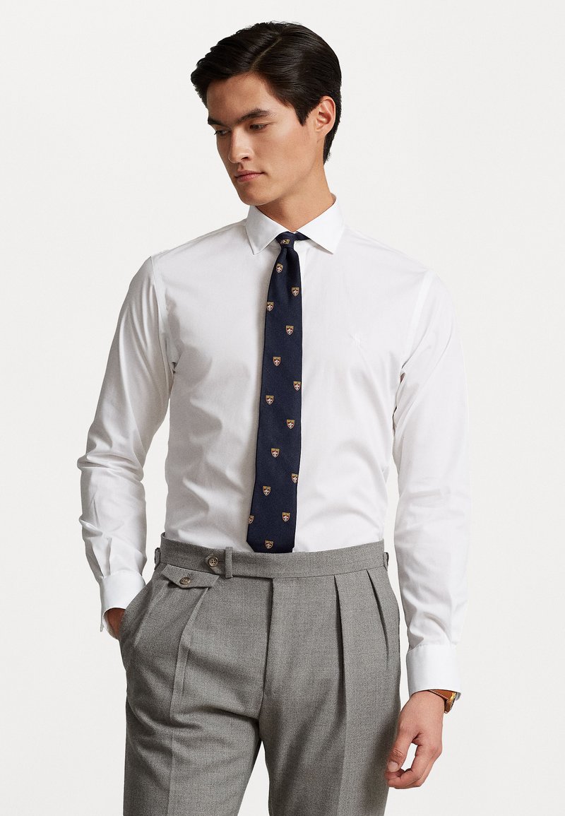 Chemise blanche à col boutonné, associée à une cravate marine à motifs de bouclier et à un pantalon plissé gris.