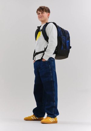 Teenager-Junge trägt weißen Pullover mit gelber Sonne, weite blaue Jeans, gelbe Turnschuhe und einen großen schwarz-blauen Rucksack, steht mit den Händen in den Taschen.