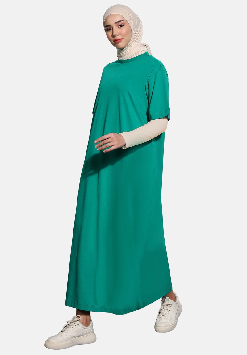 Turkoosi maxi-mekko pehmeästä kankaasta, lyhyillä hihoilla ja rentoa istuvuutta, yhdistettynä vaalean beigeen huiviin ja valkoisiin lenkkikenkkiin.