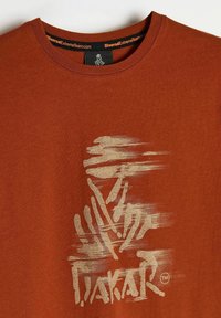 Czerwono-rdzawa, bawełniana koszulka z krótkim rękawem, z złotym graficznym wzorem przypominającym abstrakcyjne paski i dłonie, z metką przy kołnierzyku.