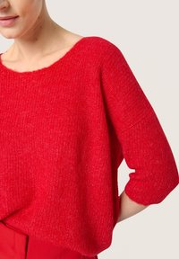 Rød strikket sweater med en bred udskæring, løs pasform og trekvartærmer. Teksturen er blød med et let fnugget udseende.