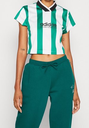 Persoon draagt een groen-wit verticaal gestreepte Adidas crop top en groene broek met trekkoord, met Adidas-logo en stergraphics.