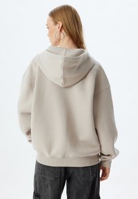 Sudadera con capucha de color beige claro, de corte relajado, con puños acanalados y una textura suave. La parte trasera presenta un diseño limpio con una capucha centrada.