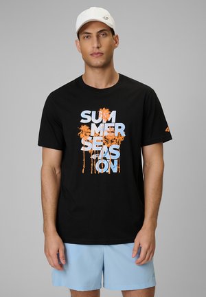 Mężczyzna w białej czapce, czarnej koszulce z napisem "SUMMER SEASON" i pomarańczowymi palmami oraz jasnoniebieskich szortach, stojący na szarym tle.