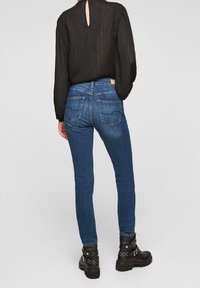 Kvinna i blå slim-fit jeans, en långärmad svart blus med nyckelhål baktill, och svarta ankelskålar med spännen och nitar.