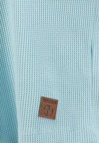 Tejido de punto waffle azul claro con una pequeña etiqueta de cuero marrón etiquetada "KEY LARGO" cosida cerca de la parte inferior.