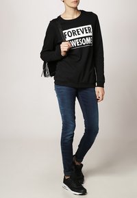 Černá mikina s nápisem "FOREVER AWESOME", modré slim-fit džíny a černé tenisky. K outfitu je přidána černá třásňová kabelka.