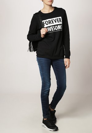 Sudadera negra con el texto "FOREVER AWESOME", jeans azul slim-fit y zapatillas negras. El conjunto se complementa con un bolso negro con flecos.