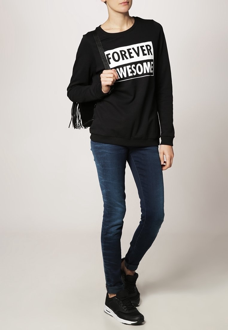 Černá mikina s nápisem "FOREVER AWESOME", modré slim-fit džíny a černé tenisky. K outfitu je přidána černá třásňová kabelka.