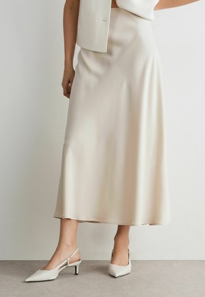 Kvinde iført cremefarvet satin midi nederdel, matchende ærmeløs blazer og cremefarvede slingback hæle med spids tå, stående op ad hvid væg.