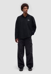 Schwarzes Longsleeve-Polo mit Kragen, ausgestattet mit einem kleinen weißen Logo. Kombiniert mit locker sitzenden schwarzen Cargo-Hosen und schwarzen Schuhen.