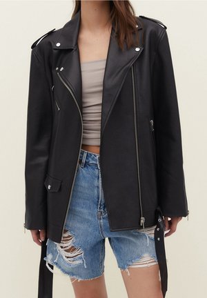 Veste de motard en cuir noir avec fermetures éclair argentées portée sur un crop top gris et un short en denim bleu déchiré.