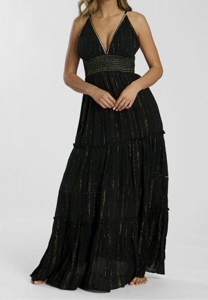 Femme portant une robe maxi noire sans manches avec des rayures verticales dorées et une taille smockée ajustée, debout pieds nus sur un fond blanc.