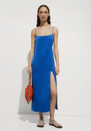 Woman in a blue sleeveless dress with thigh slit, holding a red beaded bag, wearing strappy silver sandals, standing against a plain background.