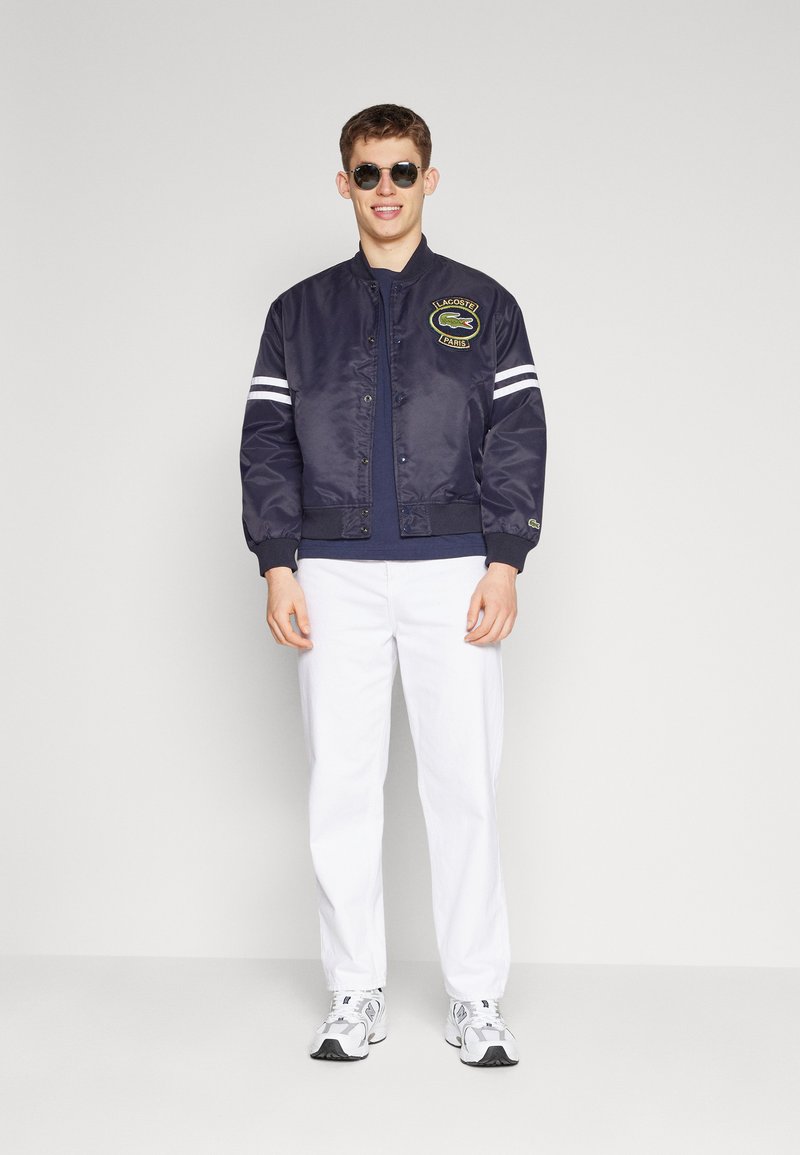 Veste de bombardier navy avec des rayures blanches, logo brodé et poignets côtelés. Associée à un pantalon blanc et des baskets grises. Modèle portant des lunettes de soleil.