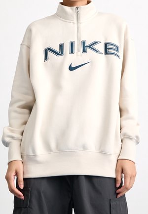 Osoba nosząca oversize beżową bluzę Nike z kołnierzem na półsupeł i czarne spodnie bojówki, stojąca na jednolitym tle.