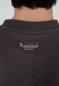 Gros plan sur le col arrière d'une chemise gris foncé avec le mot « Interdit » brodé en blanc au-dessus d'un motif à damier.