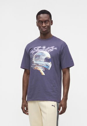 Homme portant un t-shirt violet avec un graphisme d'un casque futuriste et un pantalon Puma beige clair avec une bande latérale, debout devant un fond uni.