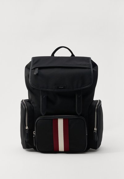 Sac à dos en nylon noir avec un rabat supérieur, comportant une poche avant zippée ornée de rayures verticales rouges et blanches et des poches latérales zippées.