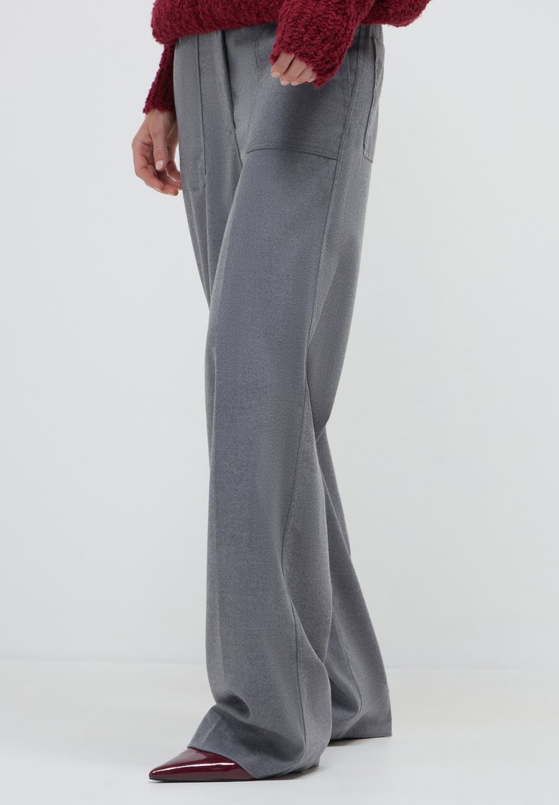 Persona che indossa pantaloni grigi larghi e scarpe rosse a punta, con un maglione rosso con texture parzialmente visibile.