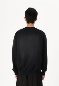 Uomo con capelli scuri in piedi di spalle, indossa un ampio sudario nero e pantaloni scuri su uno sfondo bianco semplice.