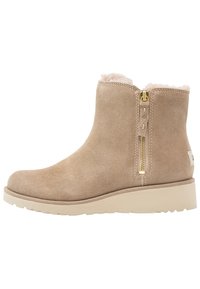 Botín de ante beige hasta el tobillo con forro de piel de crema, cremallera lateral dorada, suela de goma plana y detalles de costura sobre un fondo blanco.