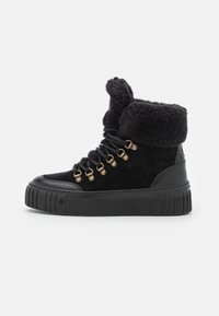 Botas de tobillo de ante negro con un puño de piel sintética negro, ojales dorados y cordones negros. La suela es texturizada y de estilo plataforma.