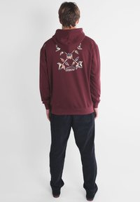 Sweat à capuche bordeaux avec un motif floral au dos, arborant un logo. Associé à un pantalon sombre, mettant en avant une coupe décontractée et une texture douce.