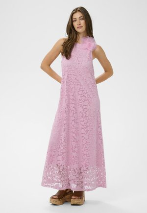 Vrouw die een roze bloemen kanten maxi-jurk zonder mouwen draagt met een grote stoffen bloem op de schouder, staand in bruine plateauzandalen.