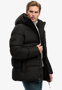 Sort dunjakke med hætte, der har lynlåslukning, trykknapper og et logo på venstre ærme. Tekstureret stof, oversized pasform.