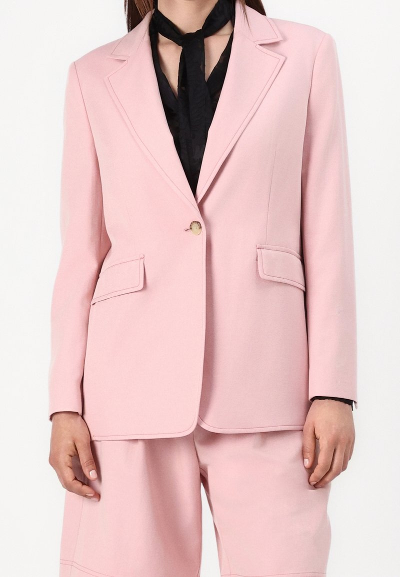 Blazer rose clair avec des revers nets, fermeture à un bouton et deux poches avant. Porté sur un haut noir avec un foulard autour du cou.