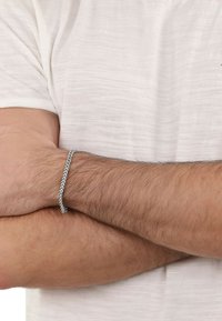 Bracciale intrecciato in argento su un polso, caratterizzato da un motivo a chevron. Il polso è parzialmente coperto da una camicia di colore chiaro e texture.