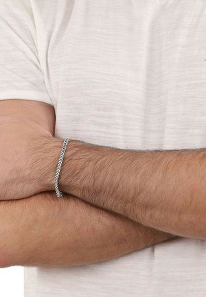 Pulsera trenzada de plata en una muñeca, con un patrón de flecha. La muñeca está parcialmente cubierta por una camisa texturizada de color claro.
