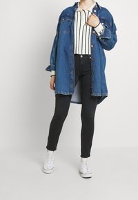 Camisa de mezclilla azul con botones naranjas, combinada con una blusa blanca a rayas y jeans ajustados negros. Zapatillas blancas completan el atuendo.