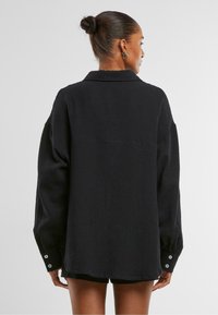 Černá oversized košile z texturované látky, vybavená límečkem, spadlými rameny a knoflíkovými manžetami. Viditelné švy na zadní straně.