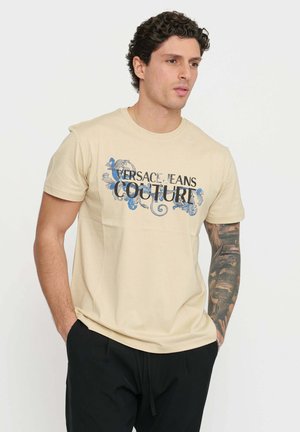 T-shirt in cotone beige con stampa floreale blu e nera e il testo "VERSACE JEANS COUTURE." Maniche corte e scollatura a girocollo.