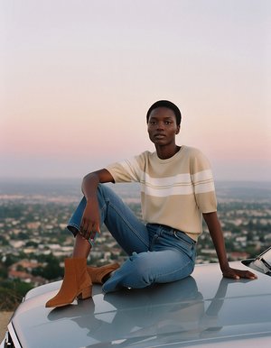 Suéter beige a rayas, jeans azules y botas de tobillo marrones. La modelo posa en el techo de un coche contra un cielo pastel con vista a la ciudad.