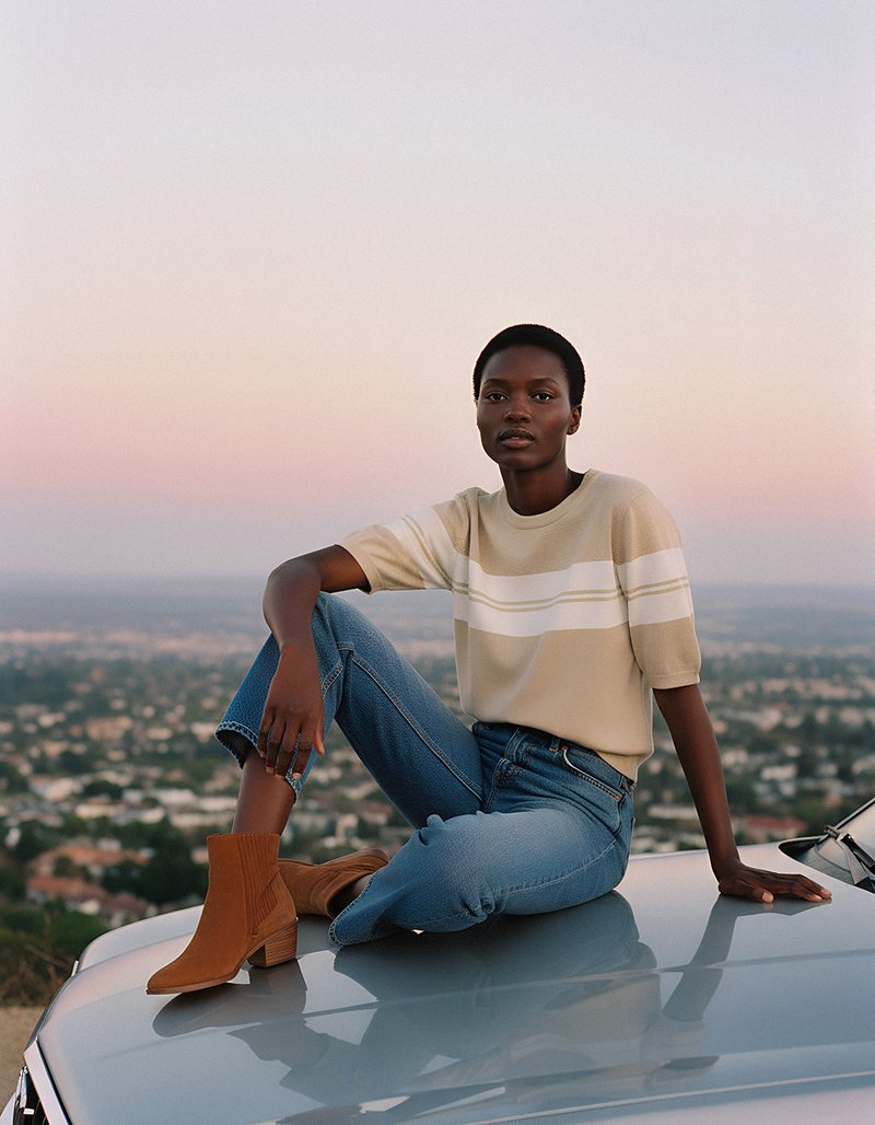 Suéter beige a rayas, jeans azules y botas de tobillo marrones. La modelo posa en el techo de un coche contra un cielo pastel con vista a la ciudad.