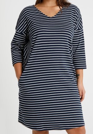 Femme portant une robe à rayures horizontales bleu marine et blanc, longueur genou, manches trois-quarts, col en V, mains partiellement dans les poches.