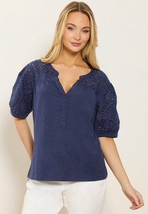 Blondynka w granatowej bluzce z ozdobnymi przelotkami na bufiastych rękawach i guzikami z przodu, w białych spodniach, stojąca na tle beżowej ściany.