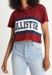 Polo shirt bordeaux a maniche corte con strisce bianche e blu navy, caratterizzato dal logo "HOLLISTER". Tessuto morbido, dettagli con bottoni.