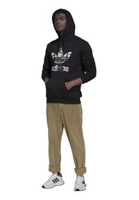 Felpa nera con cappuccio e un grande logo Adidas verde e bianco, abbinata a pantaloni di velluto a coste beige e sneakers bianche con dettagli neri.