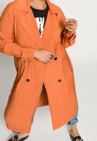 Pomarańczowy płaszcz trench z wyciętym kołnierzem, podwójnymi guzikami i bocznymi kieszeniami, nałożony na czarną koszulkę z grafiką i dżinsy.