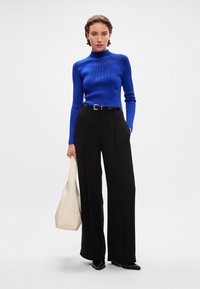 Pulôver azul de gola alta com textura canelada, combinado com calças largas pretas de cintura alta e uma mala tote bege. Cinto preto com detalhe de logotipo.