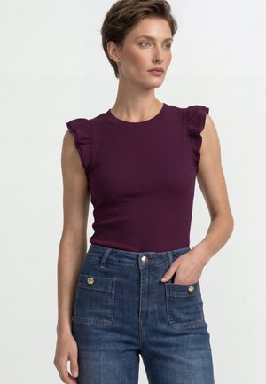 Donna che indossa un top viola senza maniche con spalle arricciate e jeans blu a vita alta, mano in tasca, guarda di lato.